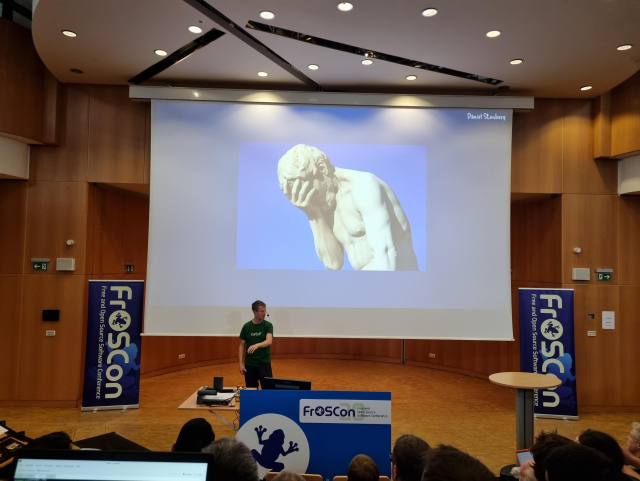 Daniel in front of a big screen showing a facepalming statue (from a talk at FrOSCon 2025)