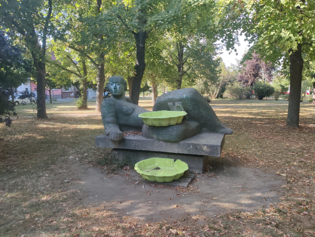 Sandstone lying figure statue, probaby woman in the middle of park. Somebody sprayed huge eyes  on it's face and added two huge plastic buckets in front of it. 