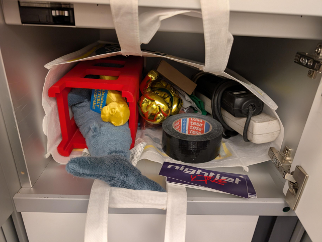 Gepäckfach in einem Nachtzug. Darin ist eine Tasche mit einer 3D-gedruckten Telefonzelle, Ottern, einer Winkekatze, einem Blåhaj und einer Rolle Gaffa. Davor liegen Sticker mit dem Aufdruck "Nightjet Ultras".