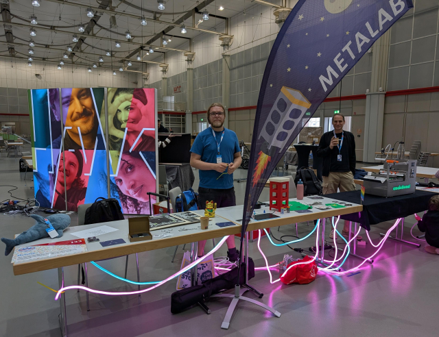 Mehrere Tische stehen nebeneinander in einer Messehalle, dahinter stehen 2 Personen, die zur Kamera schauen. Auf den beiden Tischen links liegt viel Kleinzeug, ein Plüschhai, 3D-Gedruckte Telefonzellen, Otter und eine Winkekatze. Darunter hängen Kreuz und quer pinke und blaue LED-Schlangen. Vor den Tischen steht eine Beachflag auf der Metalab steht und eine Telefonzelle abgebildet ist, die als Rakete ins All fliegt. Rechts steht ein Tisch mit einem silbernen Gerät drauf, welches mit Amalettomat beschriftet ist.