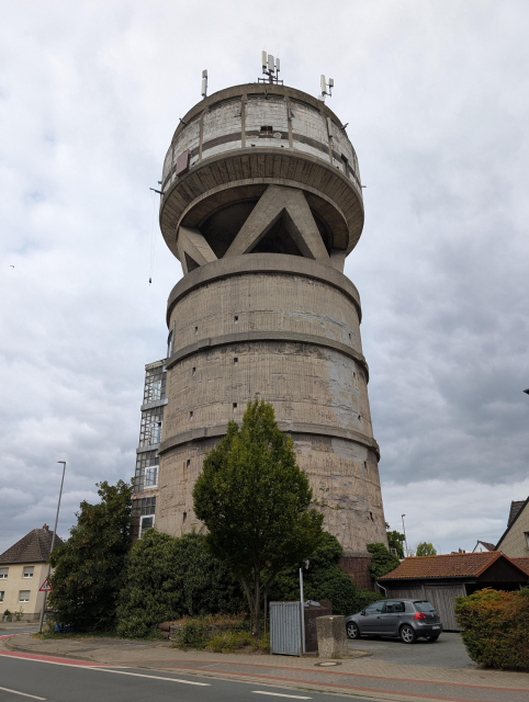 Ein runder Turm aus Beton steht neben einer Straße. Auf dem Turm steht ein weiteres rundes Gebilde mit ungefähr dem selben Durchmesser mit schrägen Betonstreben abgestützt. Am Dach sind mehrere Mobilfunkantennen. Das ganze Gebäude passt nicht zur Gegend und sieht etwas pflegebedürftig aus.