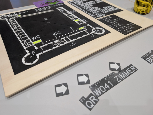 Mehrere zweifarbige Schilder aus dem 3D-Drucker liegen auf einem Tisch. Sie zeigen Text in Braille und in tastbarer Schritt sowie ein paar Pfeile. Im Hintergrund liegt ein Grundriss eines Gebäudes mit ein paar Beschriftungen und einer Legende, ebenfalls als Relief ausgeführt.