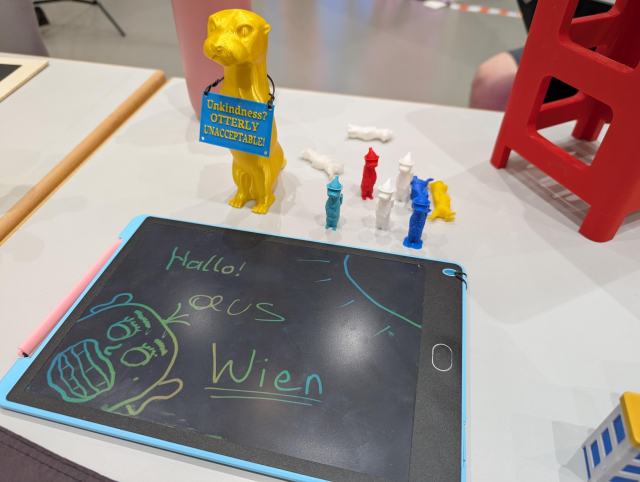 Ein Tablet mit dem Text "Hallo aus Wien" und ein aufgemaltes Gesicht. Dahinter stehen mehrere 3D-gedruckte Otter, einer mit dem Schild "Unkindness? Otterly unacceptable"