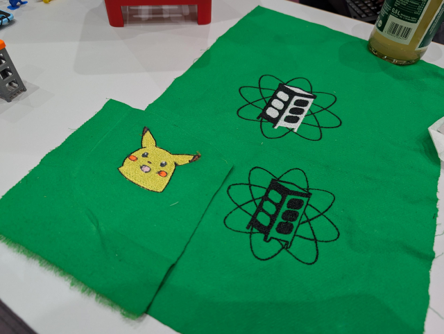 Mehrere bestickte Stoffstreifen liegen auf einem Tisch. Sie zeigen ein Surprised Pikachu und das Metalab Logo, eine Telefonzelle vor mehreren Orbitalen.