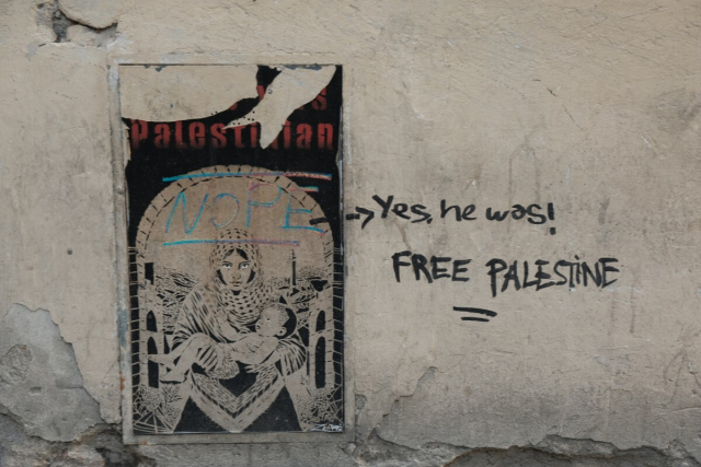 An urban, anonymous argument. A poster glued to a building's wall on the street, with "Jesus was" peeled off.  You can still see "Palestinian" and an illustration of Mary wearing a keffiyeh, carrying a baby Jesus under an arch.  The poster has been graffittied on top with NOPE.  The wall in turn has "Yes, he was! FREE PALESTINE" written next to the poster.