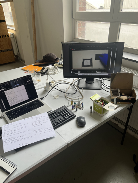 Grey desk with a laptop and external monitor showing a youtube video about how to DIY a battery. On the table are containers with various alcaline batteries and some notes on sheets of white paper.