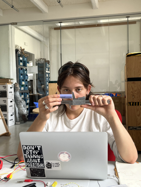 White female with brown hair measuring the size of a lithium-ion battery with a metal measuring tool and looking intently at the battery. She has a laptop in front of her.