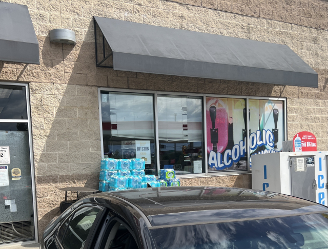 Photo of the front of a convenience store. Typical things like an ice machine bottled water, etc. A plain tan stone storefront with a dark awning over the window. 
