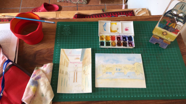 Two small sketches of Firenze, on watercolor paper, just starting to get color on them. A water container, brush, watercolor kit, and assorted trinkets on my table.