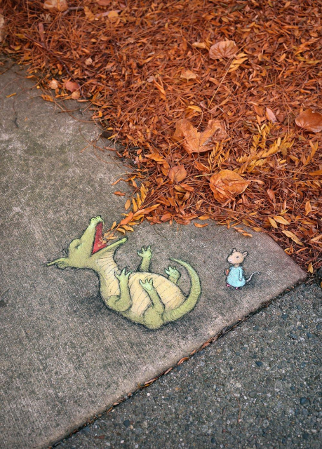 Whimsical sidewalk chalk art by David Zinn featuring a playful green dragon lying on its back, laughing as autumn leaves appear to burst from its mouth, while a small mouse in a blue dress stands nearby. The scene blends seamlessly with real fallen orange and brown leaves on the pavement, creating the illusion that the dragon is breathing autumn into life.