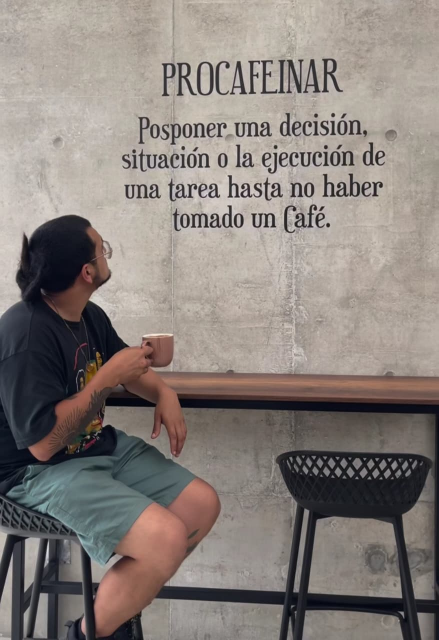 Foto de una persona sentada en un banco en una cafetería, está viendo hacia la pared en donde está escrito: "PROCAFEINAR.
Posponer una decisión, situación o la ejecución de una tarea hasta no haber tomado un Café"