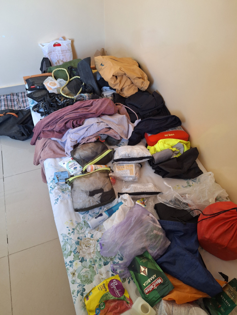 Ein simples Bett in einem kargen Zimmer, voll mit unordentlich Sachen und Dingen. 