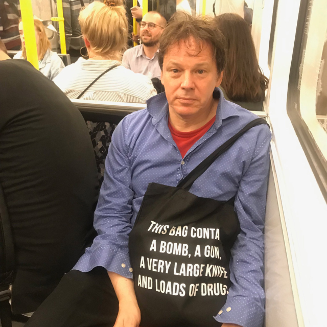 
A photograph of David Graeber carrying a bag on the subway. The bag reads "This bag contains a bomb, a gun, a very large knife, and loads of drugs."
