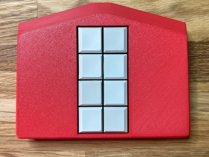 Mobile Braille keyboard Metabraille with 8 keys. The case is red and the buttons are white. The case is about pentagonal. A rectangle is extended by a trapezoid on the broad side. The shape resembles a house with windows in the middle.