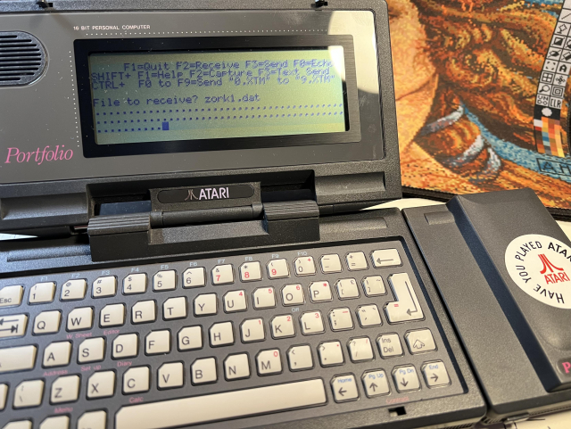 An Atari Portfolio computer with a serial interface. On the screen is a terminal program XTerm downloading "zork1.dat" showing progress as dots.
