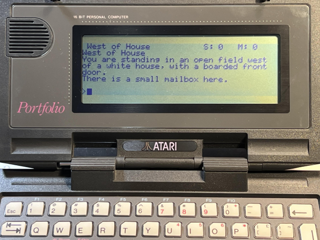 An Atari Portfolio running Zork 1. On the monochrome screen it says "West of House. You are standing in an open field west of a white house, with a boarded front door. There is a small mailbox here." There's a > prompt and a flashing cursor at the bottom of the screen.