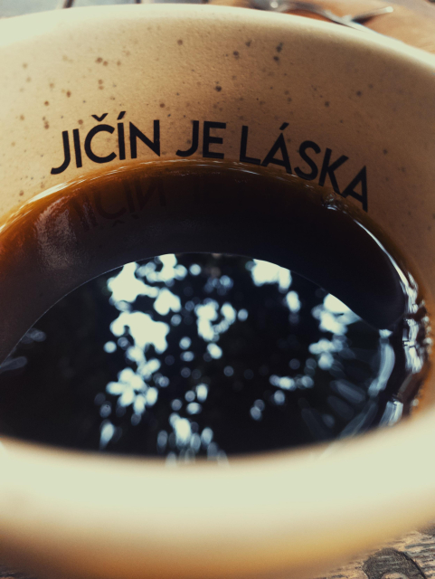 Detailní pohled na hrnek s kávou s nápisem „JIČÍN JE LÁSKA.“ Hrnek je naplněný tmavou kávou a na hladině jsou viditelné odlesky světla z okolních stromů.