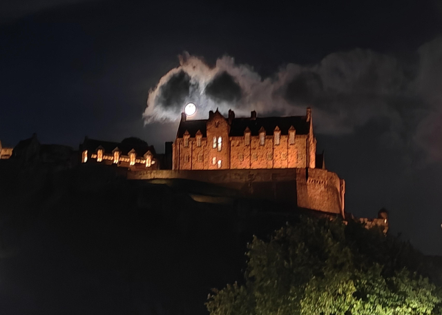 A colour photograph of the moon coming out from behind thick cloud, behind Edinburgh Castle. 

The castle, in the centre of the picture, is illuminated and appears bathed in orange-red light. 

The moon is just above the castle, and is the brightest thing in the picture. The moon is a white disc, with no detail visible.

It is the clouds which make the picture. There are two dark circles of cloud, one on each side of the moon, which is lighting up their edges so they look like eyes or spectacles. 

The rest of the sky is very dark, and the cliffs of Castle Hill are also dark. 

In the bottom right corner the top of a tree can be seen, lit up by a street lamp. The leaves are bright green.