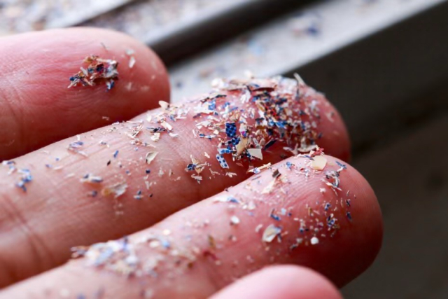 A close-up of a hand covered in colorful, fine shavings or flakes (microplastics), with a blurred background suggesting a workspace.