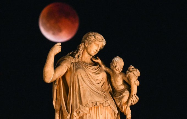 A nighttime photograph of a classical stone statue set against a pitch-black sky. The statue depicts a woman in draped robes and a small child holding a cornucopia. Positioned just above the woman's raised hand is a large, deep-red "Blood Moon," captured during a total lunar eclipse.