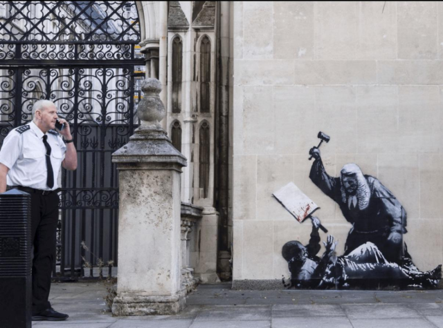 A photograph of an intricate stencil in black, white, and gray, depicting a life-size protestor being beaten bloody on the pavement by a bewigged judge with a gavel. The stencil appears to be on the gray stone facade of a building with a filigree iron gate and an annoyed-looking functionary on his mobile.