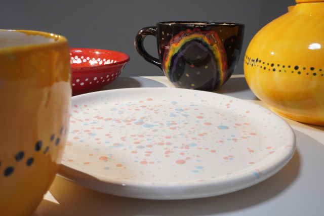 Mehrere handgemachte Keramikstücke stehen auf einem weißen Tisch. Vorne liegt ein flacher Teller mit pastellfarbenen Sprenkeln in Rosa und Hellblau. Links ist eine gelb glasierte Schale mit einem schwarzen Punktmuster zu sehen, rechts daneben ein bauchiges Gefäß in derselben Farbgestaltung. Dahinter stehen eine schwarze Tasse mit einem bunten Regenbogenmotiv und eine rote Schale mit weißen Punkten am Rand.