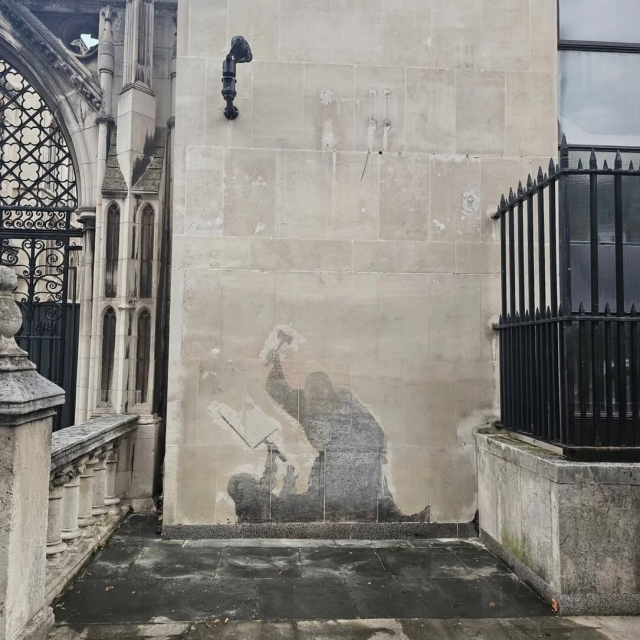 A view from the street, showing one entrance to the Royal Courts of Justice in London, off to the left side; the center of the photograph is the wall where a Banksy has been erased, leaving behind the black and white silhouette of a bewigged judge, holding a gavel like a hammer over their head while standing over a prostrate protester, who is unarmed, holding up one empty hand and a placard on the other hand. The stain is very clear against the rest of the wall.