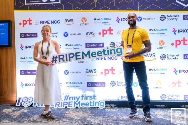Two smiling participants pose in front of a sponsor backdrop at RIPE 90. The person on the left wears a white dress and holds a large cutout reading “#RIPMeeting,” while the person on the right, in a yellow shirt and dark jeans, also holds the sign. Another cutout on the floor reads “#myfirstRIPMeeting.” The backdrop displays logos of RIPE NCC, AWS, Internet Society, Verisign, and other event sponsors.
