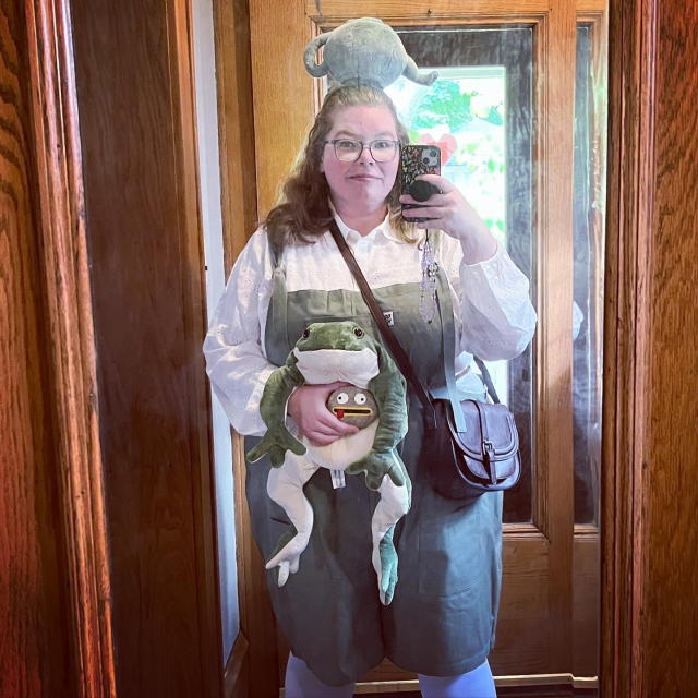 Ashe dressed up as Greg from Over the Garden Wall. She is wearing green short overalls, a white shirt, and an upside down plush teapot on her head. She has a brown leather purse worn across her body and in her hands are a plush frog and a rock with a silly painted face on it.