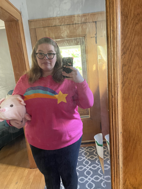 Ashe dressed as Mabel from Gravity Falls. She is wearing a pink sweatshirt with a rainbow shooting star on it and a pink headband. She is holding a plush pig.