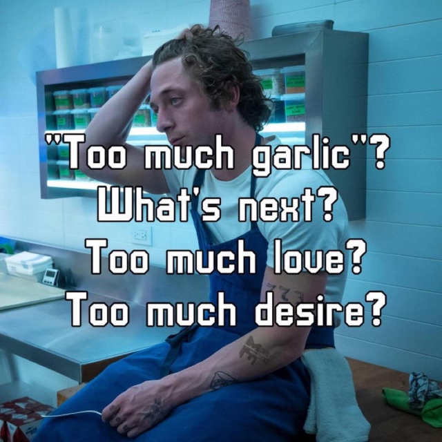 Bear, from the TV series The Bear with curly hair and visible forearm tattoos sits on a stainless steel kitchen counter, wearing a white shirt and blue apron. They rest one hand on their head, looking contemplative or exasperated. Behind them are shelves with kitchen supplies and a paper towel roll. Overlaid text reads:  
"Too much garlic"? What's next? Too much love? Too much desire?