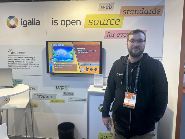Close up photo of Igalia's booth at IBC 2025. In the foreground right is Thibault Saunier standing up facing to the camera with a welcome smile. In the background a wall decorated with "Igalia is open source", and additional messages  that combines with "Igalia is", such as "web, standards, and for everyone". In the background at the middle there is a TV demoing a game in Amazon Luna using GStreamer WebRTC on WPE.