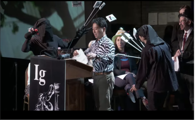 Scientist having just taken his shirt off to reveal a zebra print  is surrounded by frustrated "flies" held by assistants wearing black.