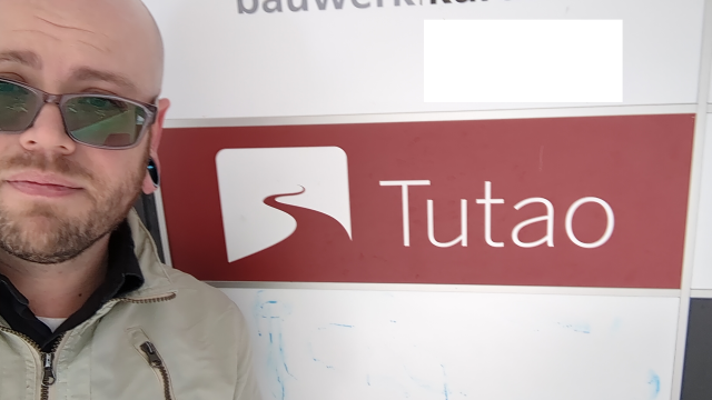 A selfie of me (bald white man with a short beard, glasses, tan jacket) standing next to the sign for Tutao on the building where their office is located.