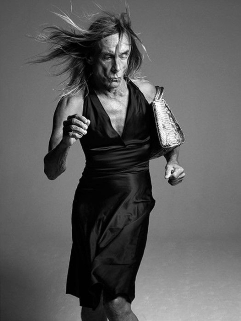 Black-and-white photo of Iggy Pop in motion, captured mid-stride as he runs forward wearing a sleeveless dress. His long hair blows dramatically backward, and he clutches a handbag. 
