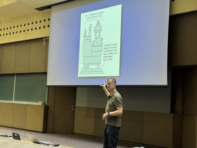 “Badger” on stage pointing to the XKCD small-one-developer-tool-holding-up-the-industry cartoon on the screen behind him.