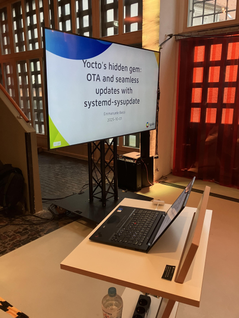A podium with an open laptop on it; behind it, a large display with a presentation titled “Yocto’s hidden gem: OTA and seamless updates with systemd-sysupdate”