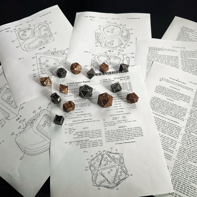 Photograph of acrylic and metal 7 piece braille rpg sets atop scattered pieces of paper. The documents are details of the US Patent 12,415,127 and include pages with illustrations of the DOTS RPG Project braille dice having specific areas of the dice tagged with reference numbers and multiple pages of text explaining the unique design and function of each element of a die. 
