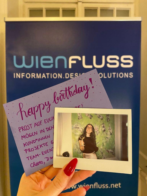 Daniela holding a Polaroid picture of herself together with a birthday note in front of a Wienfluss company roll-up.
