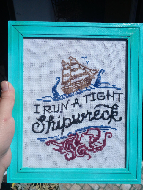 An embroidery piece. A wooden ship is attacked by a bordeaux giant octopus. Through the middle the phrase "I run a tight Shipwreck" in black on a wavy pattern.