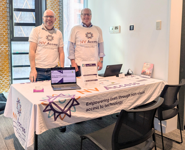 Quentin and James standing behind the NV Access table at Tech Fest with a laptop setup and brochures