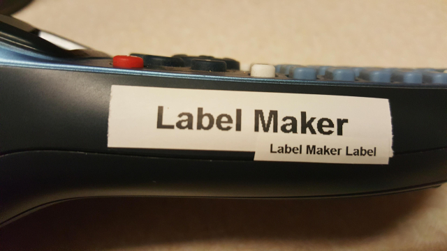 A label on the side of a handheld label maker: black text on white tape, the label reads: "Label Maker". On the lower right of the label is a smaller label, with a smaller font, which reads "Label Maker Label".