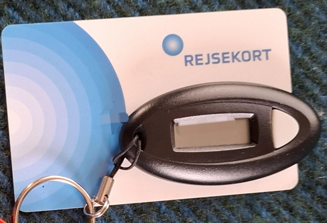 A physical travel card, credit-card sized. White with blue logo and the name Rejsekort (literally, Danish, 'Travel Card') on it. On top of it, a black oval fob with a display window and a grey button beside it. You click the button to get the 6-digit  code you need to verify logging on to bank services, government services, your digital postbox 'Ebox' etc in Denmark.
