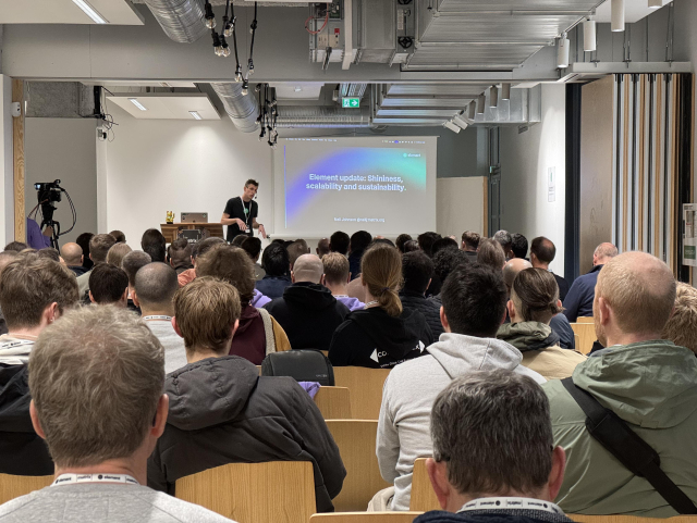 A room full of people and a speaker in front of their slides spelling out “Element update: Shininess scalability and usability.”