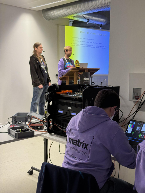 A picture of two young people on stage talking to the audience. In the foreground, a volunteer with a matrix hoodies is monitoring a computer displaying a picture of the speakers