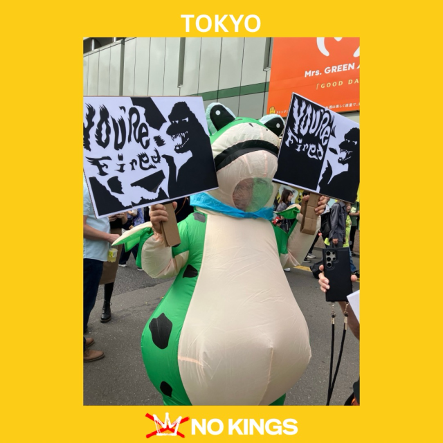 A photo of a person in a frog costume holding sings that read “You’re Fired” (alongside a drawing of Godzilla) at the No Tyrants protest in Tokyo, Japan.