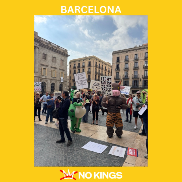 A photo of over a dozen people at the No Tyrants protest in Barcelona, Spain. A person in a frog costume is holding a sign that reads “I STAND WITH OREGON/EL PUEBLO UNIDO/ABOLISH ICE/NO TYRANTS.”