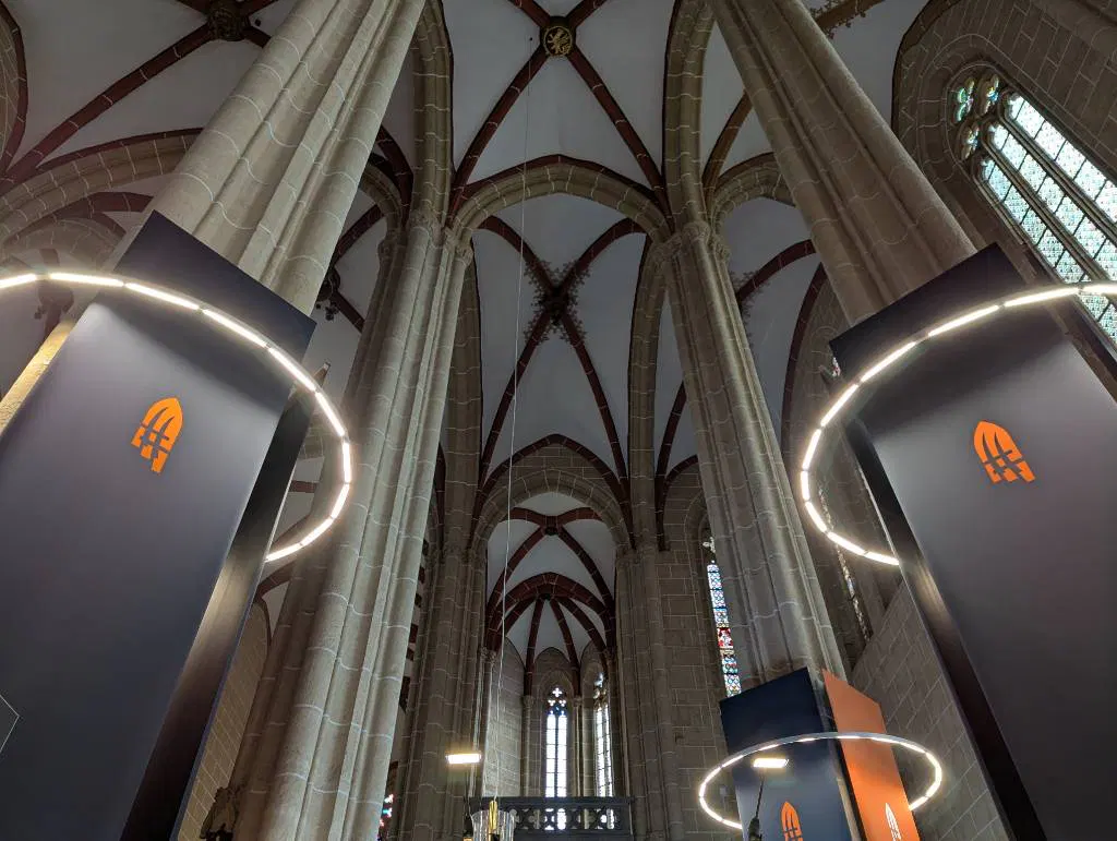 Drei Säulen der Marienkirche gegen die Gewölbedecke fotografiert. Sie umgeben einen Lichtring, an ihnen sind große Transparente angebracht.

Darauf das Logo der Landesausstellung, bestehend aus drei stilisierten Spitzen von Hellebarden, die zusammengefügt wie ein Kirchenfenster aussehen.