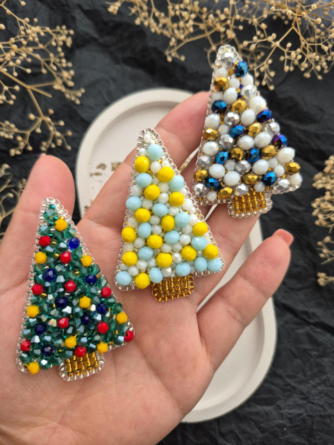 Close-up shot: an open palm holds three handmade Christmas tree-shaped brooches. The brooches are densely embroidered with beads and crystals: 1) green with red, yellow, and blue elements; 2) yellow-blue with white beads; 3) white-gold with blue and silver crystals.