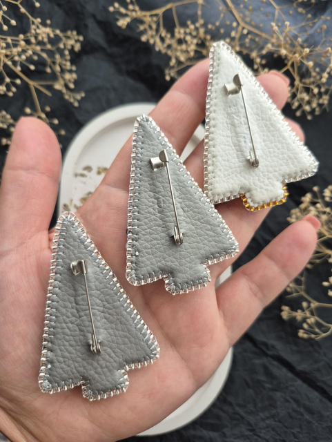 Close-up shot: An open palm holds three Christmas tree-shaped brooches, turned inside out. Each brooch has a metal pin fastening and a faux leather trim (two gray, one white). A neat beaded edge is visible.